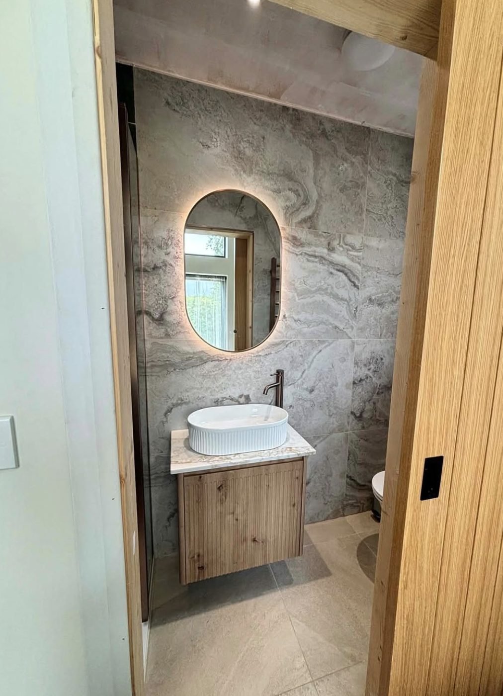 Powder room with a fluted basin on a timber vanity, oval backlit mirror and marble effect tiling